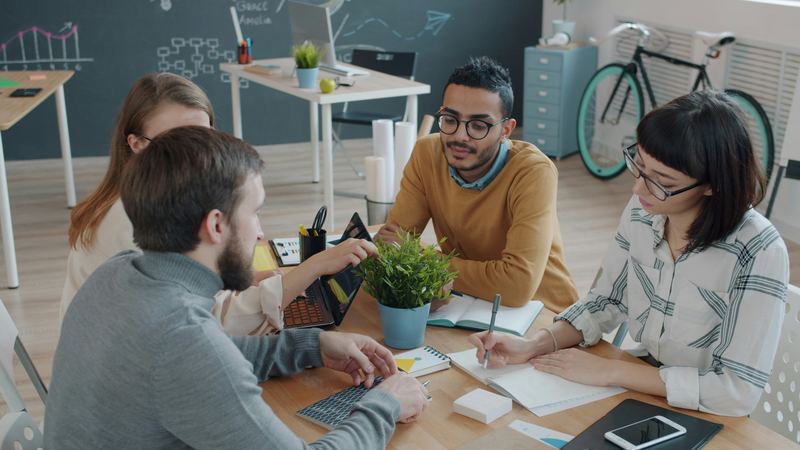 Four colleagues gathered around a table with notebooks, a laptop and a small plant in a bright office