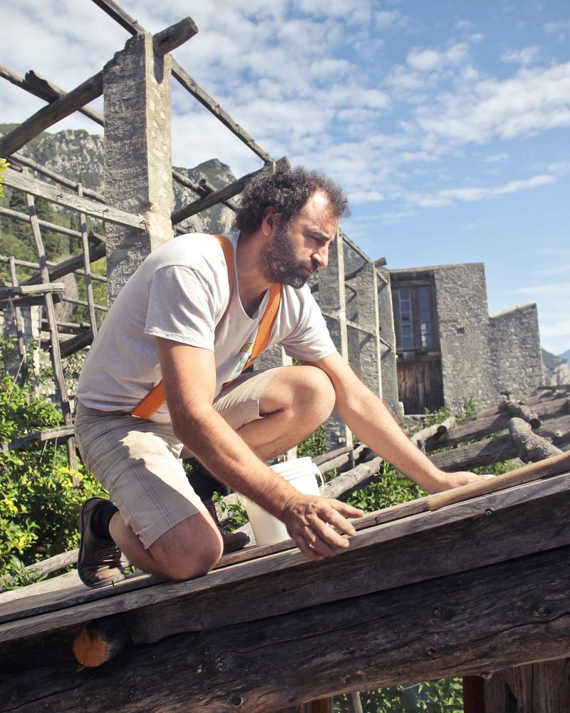 A man in a light T-shirt crouching on a wooden roof frame, working with a hand tool